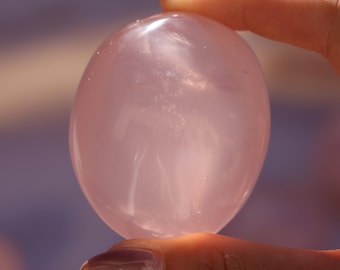 Può includere: Un cristallo di quarzo rosa liscio di forma ovale, di colore rosa traslucido. Il cristallo è tenuto in una mano.