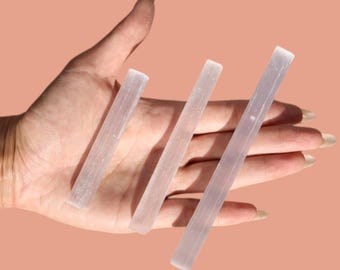 May include: Three translucent, rectangular selenite crystal wands of varying lengths are displayed. The wands are held in a hand against a peach-colored background. The crystals have a soft, glowing appearance.