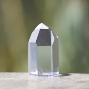 May include: A clear quartz crystal point with a faceted design. The crystal is transparent, allowing light to pass through, and sits on a light-colored surface. The background is blurred, with shades of green and brown.