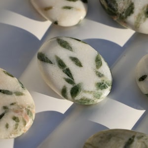 May include: Close-up of a white oval-shaped stone with green flecks. The stone is smooth and polished.