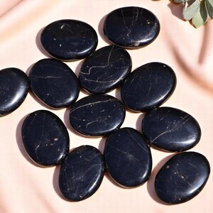 May include: A collection of polished, oval-shaped black stones with subtle white veining. The stones are arranged on a light pink surface, creating a visually appealing display. The smooth, dark surfaces of the stones reflect light, enhancing their natural beauty.