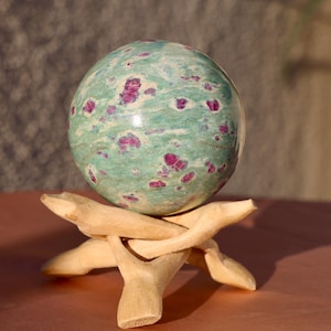 Peut inclure: Une sphère de rubis en zoïsite verte et rose sur un support en bois. La sphère est polie et présente un motif tourbillonnant de vert et de rose.