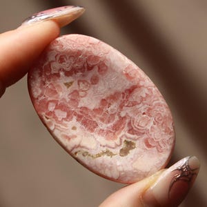 May include: A polished, oval-shaped gemstone with a swirling pattern of pink and white hues. The stone has a smooth surface and is held between two fingers. The gemstone's color is a mix of light and dark pink, with some areas appearing almost white.