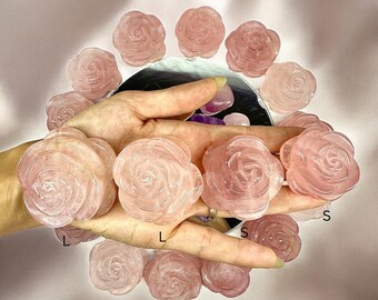 May include: A selection of rose quartz carvings in the shape of roses. The carvings are in different shades of pink and are arranged around a reflective surface. The image is taken from above, highlighting the detailed craftsmanship of the carved flowers.