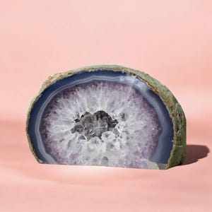 May include: A large, half-round slice of agate with a purple and white banded pattern. The agate has a natural, rough edge and a smooth, polished interior. The centre of the agate is filled with clear quartz crystals.