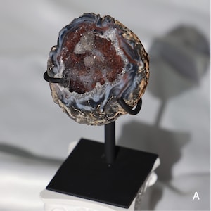 May include: A geode with a brown, white, and gray exterior. The geode is cut open to reveal a crystal-filled interior. The geode is displayed on a black metal stand.