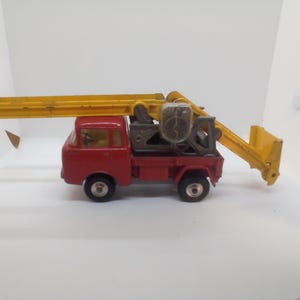 May include: A vintage red toy truck with a yellow crane arm and bucket. The truck has black wheels and a detailed crane mechanism. The crane arm extends horizontally, and the bucket is angled downward.