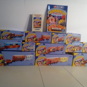 May include: A collection of Corgi Chipperfields Circus toy trucks and trailers in their original boxes. The boxes are blue and yellow, featuring circus-themed graphics and the Corgi logo. A collector's album and a circus poster are also present.