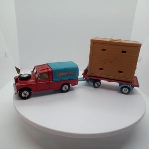 May include: A red toy truck with a blue cover and a trailer. The truck has black wheels and a spare tire. The trailer is brown and has a rectangular shape. The truck is connected to the trailer.