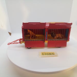May include: A red toy circus wagon with two compartments, each housing a lion. The wagon has a yellow roof and a red tow bar. The word "LIONS" is printed on a small yellow sign.
