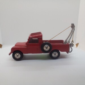 May include: A vintage red tow truck toy with black tires and a spare tire. The tow truck has a silver tow arm and a hook. The toy is a classic collectible item.