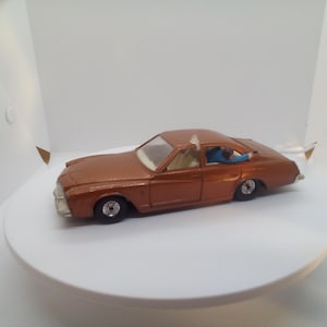 May include: A copper-colored toy car with black wheels and a white interior. The car has a driver figure visible inside. The car is on a white surface.