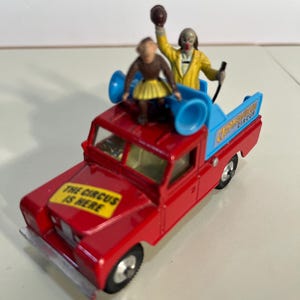 May include: A red toy truck with a blue bed, carrying a clown and a small figure. The truck has the text "THE CIRCUS IS HERE" on the hood and "CHAPMAN BROS CIRCUS" on the bed. The clown is wearing a yellow jacket.