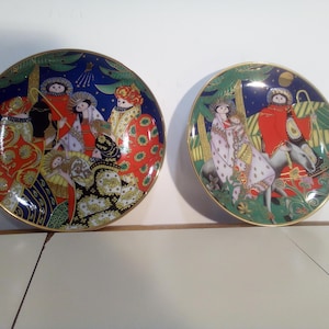 May include: Two decorative ceramic plates with gold rims. One plate features a group of figures in ornate robes against a dark blue background. The other plate depicts figures on a donkey with a green and blue background.