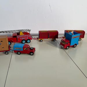 May include: A collection of vintage toy trucks and trailers, including a red fire truck with a ladder, a red truck with a blue top, and several red trailers. The trucks are labeled "Chipperfields Circus".