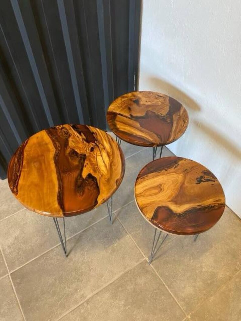 May include: Three round wooden tables with black metal hairpin legs. The tabletops are made of wood with a natural wood grain and a brown, orange, and red epoxy resin finish.