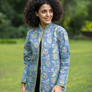 以下が含まれることがあります： 緑と紫の花柄の青いキルティングジャケット。 ジャケットはマンダリンカラーで、ライムグリーンのトリムが付いています。 袖は長く、ジャケットは前面が開いています。 背景はぼやけた緑色です。