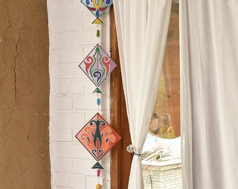 May include: A decorative hanging mobile featuring three diamond-shaped ornaments with colorful, intricate designs. The ornaments are suspended by a string with beads and a tassel, hanging against a white brick wall and a sheer white curtain.