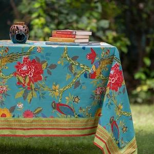 May include: A teal tablecloth with a floral and bird print. The tablecloth has a red, green and gold border. The tablecloth is laid out on a green grassy surface with a stack of books and a ceramic vase on top.