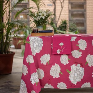 May include: A pink tablecloth with a white floral pattern. The tablecloth is laid out on a table with a wooden tray holding glasses and a small vase with white flowers.