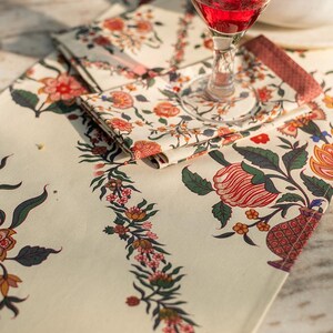 May include: A white table runner with a floral pattern in shades of red, orange, green, and blue. The runner features a repeating border of vines and flowers. A glass of red wine and a white bowl are on the table runner.