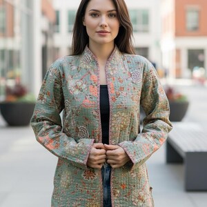 Puede incluir: Chaqueta acolchada verde claro con estampado floral en tonos naranja, marrón y rosa. La chaqueta tiene cuello mandarín y ribete rosa. La chaqueta se lleva abierta, revelando una camiseta negra. La chaqueta mide unos 76 cm de largo.