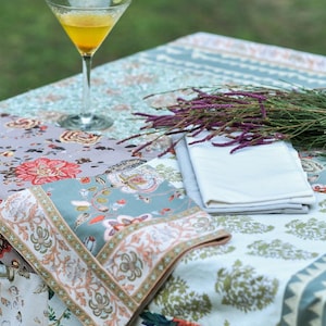 May include: A light blue tablecloth with a floral pattern and a white napkin. A glass of yellow liquid with a lime wedge sits on the tablecloth. A bouquet of purple flowers sits on the tablecloth.