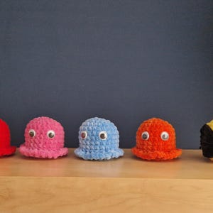 May include: A row of five handmade crochet figures, inspired by the classic arcade game Pac-Man. The figures include four colorful ghosts in red, pink, blue, and orange, and a yellow Pac-Man character. Each ghost has googly eyes.