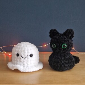 May include: A white crocheted ghost figure with black eyes and a stitched smile positioned next to a black crocheted cat figure with bright green eyes. Both plush items are crafted from a soft, textured yarn material and sit side by side.