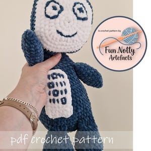 May include: A handmade blue and white crochet doll with a smiling face and a rectangular design on its front. The doll has a soft, plush texture and is labeled "pdf crochet pattern" with the Fun.Notty Artefacts logo.
