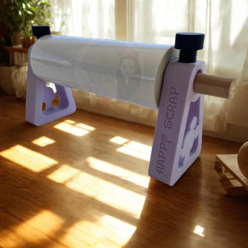 May include: A light purple craft paper dispenser with the text "HAPPY SCRAP" on the side. The dispenser holds a roll of clear paper with a cartoon character design. The dispenser is on a wooden surface.
