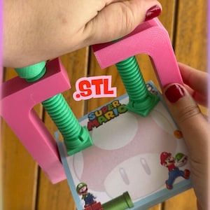 May include: A pink 3D-printed clamp holding a Super Mario-themed notepad. The clamp has green screw mechanisms and the notepad features Mario characters and a mushroom design. The word "STL" is printed on the image.