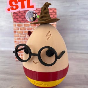May include: A novelty egg-shaped figurine with a brown Sorting Hat, black round glasses, and a lightning bolt scar. The egg is tan with a red and yellow striped base, and the word "Gryffindor" is printed on the base.