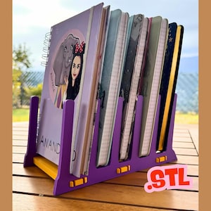 Puede incluir: Un organizador de cuadernos morado y amarillo que contiene varios cuadernos. El organizador tiene un diseño ranurado y la palabra "ANAND" es visible en uno de los cuadernos. Los cuadernos tienen varias portadas con ilustraciones y diseños.