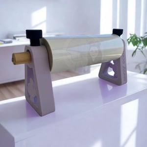 May include: A light gray paper roll dispenser with a roll of clear paper. The dispenser has a wooden dowel and black screw knobs. The supports are light gray with a decorative cutout. The text "HAPPY SCRAP" is visible on the side.