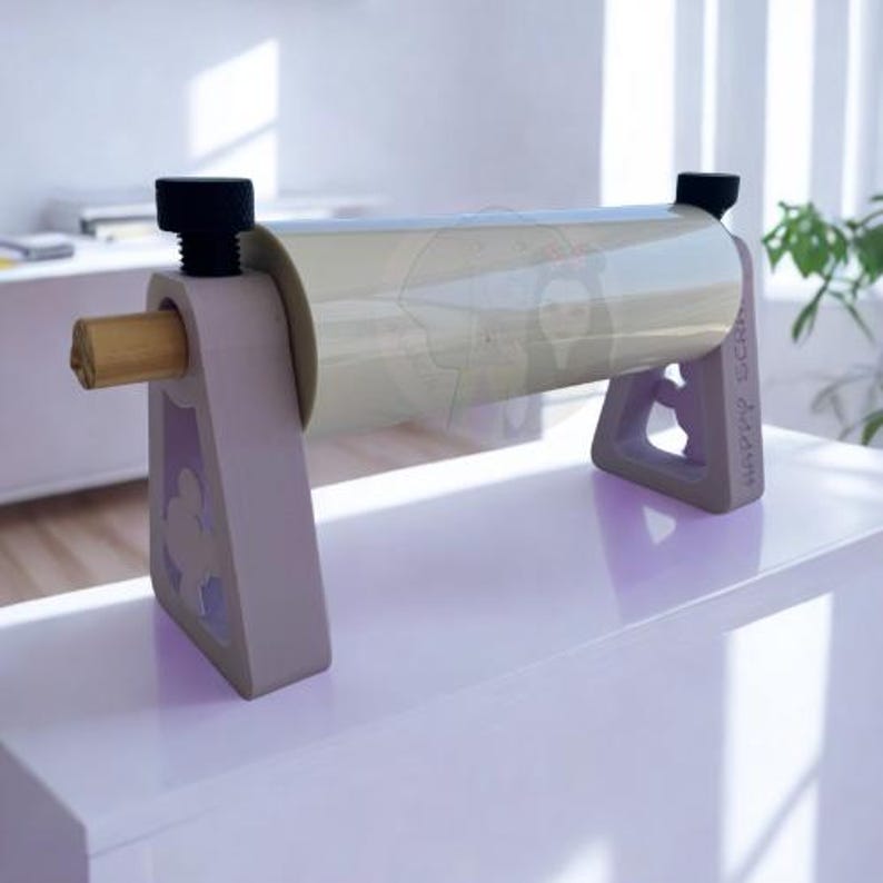 May include: A light gray paper roll dispenser with a roll of clear paper. The dispenser has a wooden dowel and black screw knobs. The supports are light gray with a decorative cutout. The text "HAPPY SCRAP" is visible on the side.