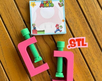 May include: Two pink clamps with green screw mechanisms sit next to a Super Mario themed notepad. The notepad features a large mushroom design and the words "Super Mario". A small sticker with the letters "STL" is also present.