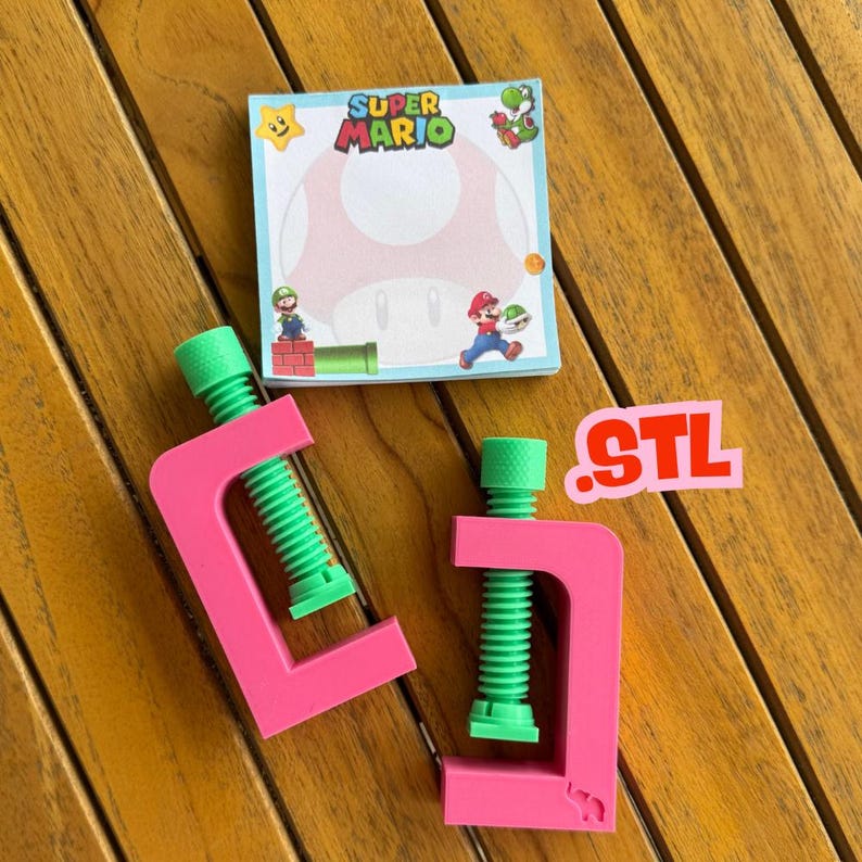 May include: Two pink clamps with green screw mechanisms sit next to a Super Mario themed notepad. The notepad features a large mushroom design and the words "Super Mario". A small sticker with the letters "STL" is also present.