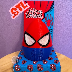 May include: A red, blue, and black Spider-Man-themed Easter egg. The egg is on a blue box with a Spider-Man pattern. The background features a red and blue panel with a spider web design and the words "Felices Pascuas".