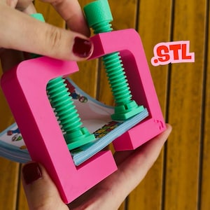 May include: A pink clamp with green screw mechanisms holding a stack of colorful papers. The clamp has the letters "STL" in the upper right corner. The clamp is held by a person with red nail polish.