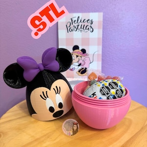 May include: A pink plastic Easter egg with a Minnie Mouse head on top. The egg is open and filled with candy and a chocolate egg. The text "Felices Pascuas" is visible on a card in the background.