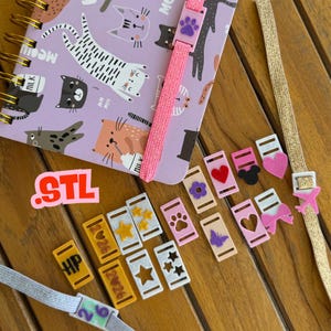 May include: A spiral-bound notebook with a cat pattern, a pink elastic band, and various decorative bookmark charms. Charms include hearts, stars, paw prints, and a butterfly. The word "STL" is also visible.