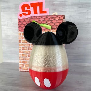 Puede incluir: Un contenedor en forma de huevo de Mickey Mouse impreso en 3D con orejas negras, una parte superior negra, una sección central blanca y una parte inferior roja con círculos blancos. El contenedor está sobre una superficie de madera.