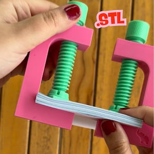 May include: A pink and green clamp holding a stack of white paper. The clamp has a screw mechanism with a green threaded rod and a green top. The text "STL" is visible in the upper right corner.