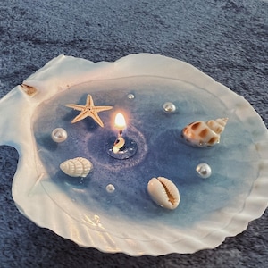 May include: A white seashell with a blue and white resin interior. The resin contains a small candle, a starfish, and several seashells.