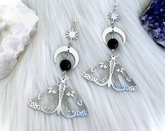 May include: A pair of silver-colored dangle earrings. Each earring features a moth design, a crescent moon, a star, and a black bead. The earrings are displayed on a white, fluffy surface.