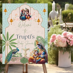 May include: A welcome sign for a baby shower with a blue and green floral design. The sign features a peacock, a fountain, and a family of three figures. The text on the sign reads "Welcome to Trupti's Baby Shower".
