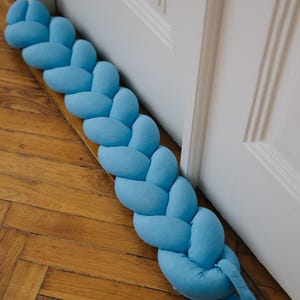 May include: A light blue braided doorstop with a loop at the end. The doorstop is made of fabric and is lying on a wooden floor.