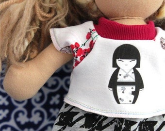 May include: A white doll with long brown hair wearing a white shirt with red trim, a black and white patterned skirt, and a red and white floral print on the sleeve. The shirt has a graphic of a Japanese doll wearing a kimono. The text "Doll Clothing by Lil Blue Boo" is printed on the bottom of the image.