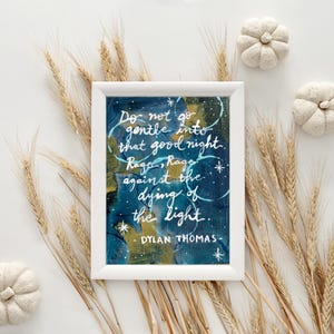 May include: White framed art print with the quote "Do not go gentle into that good night" by Dylan Thomas. The artwork features a dark blue and gold abstract background with white lettering. Decorative wheat stalks and small white pumpkins surround the framed print.